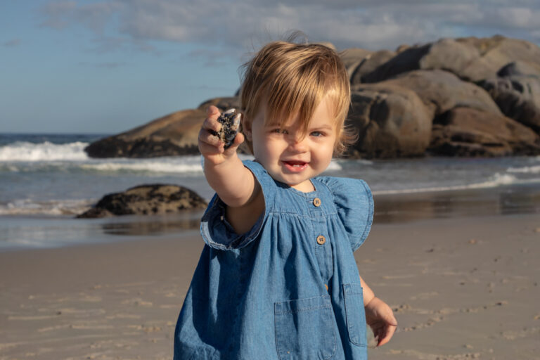 Toddler girl holding out sea shell on the beach by Katja Photography